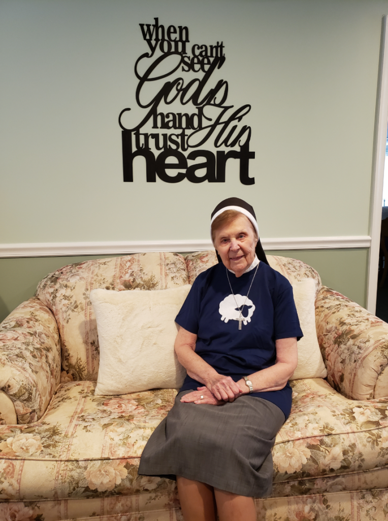 Sr. Eunice, a nun sitting on the couch and smiling wearing a flocknote shirt